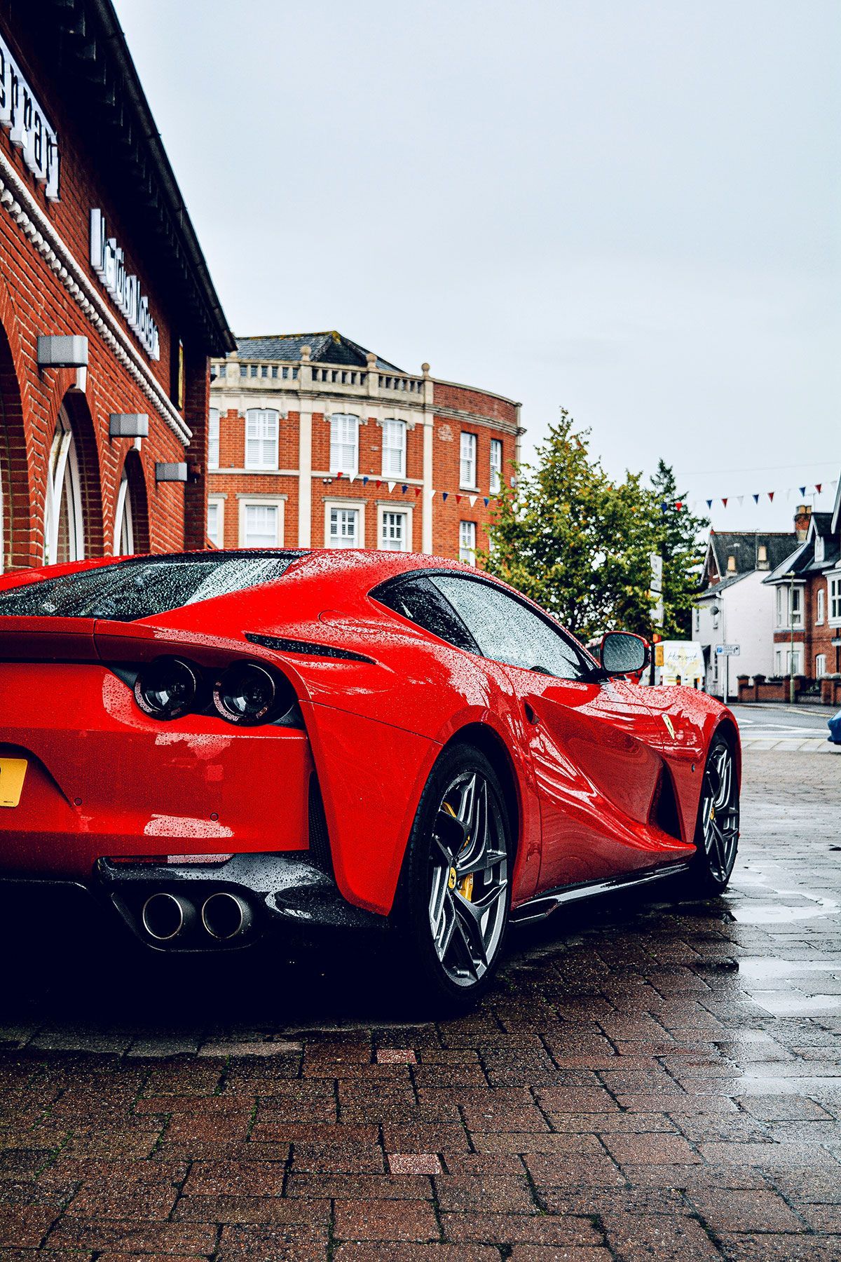 Red sports car parked on wet brick road near a brick building.